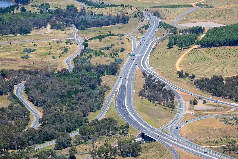 Aerial view of a multi-lane highway interchange in Canberra, with curved on-ramps and off-ramps cutting through dry grassland, scattered trees, and light traffic moving in both directions.